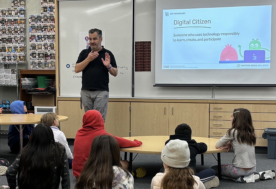 male teacher stands at white board that has the words Digital Citizen as several students sit on the floor listening to him