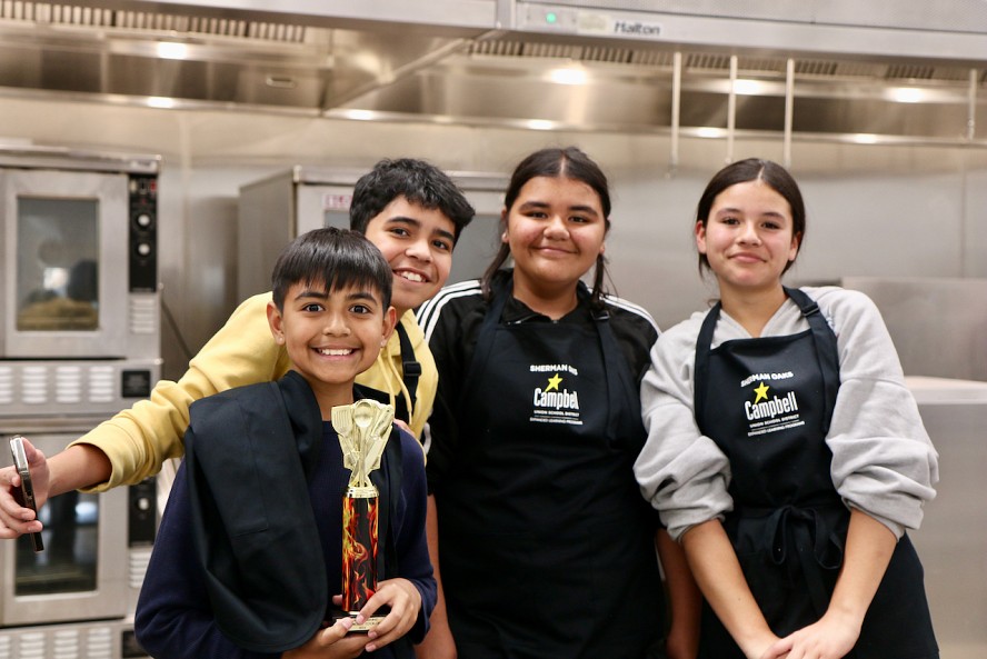 4 middle school students (2 boys and 2 girls) standing together and smiling as one boy holds a winners trophy