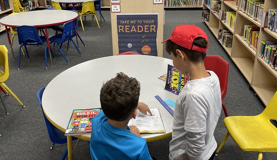 two boys in library in front of a sign that says Take Me To Your Reader
