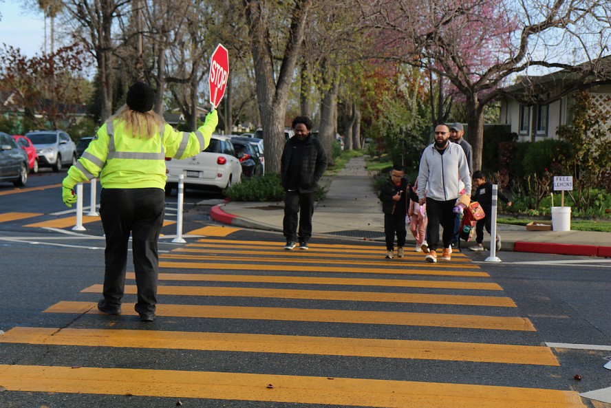 woman crossing guard holds a stop sign as a couple of children and adults crosse the street marked with wide yellow lines.