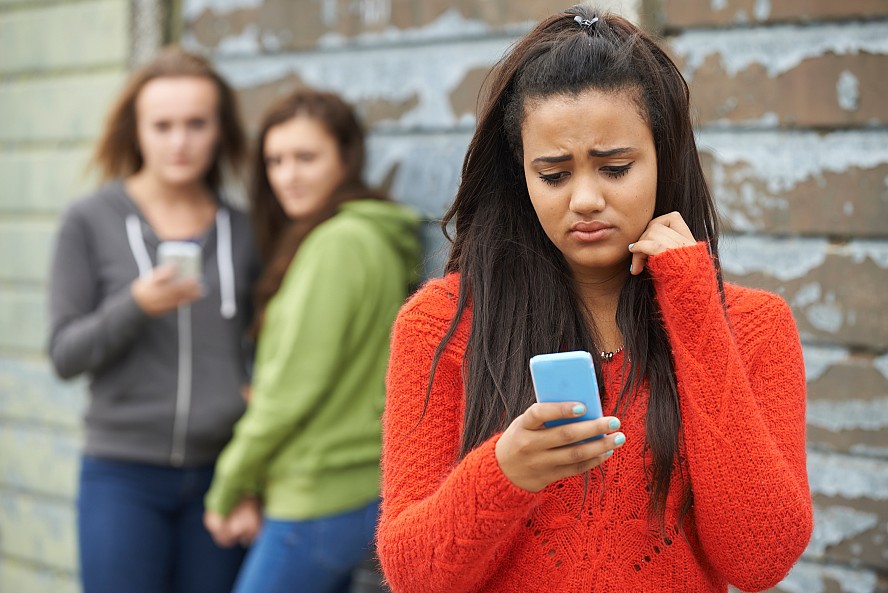 unhappy adolescent girl looking at her phone as two others behind her look and laugh at her