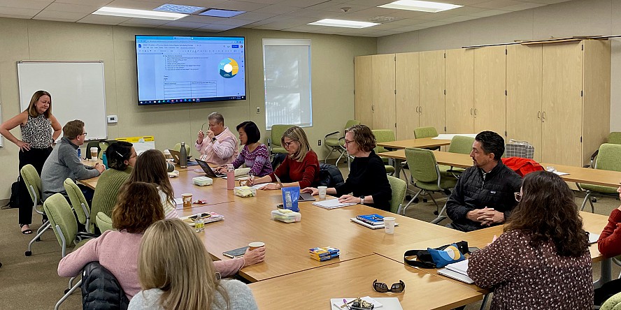 men and women at a meeting table as a facilitator stands by and a presentation is projected on a screen