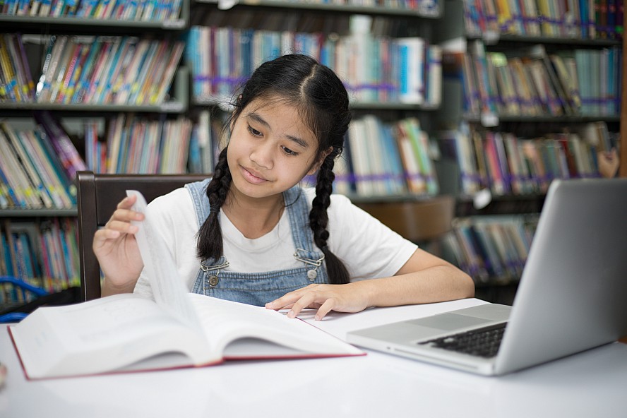 adolescent girl sitting at a table in a library is looking at a book and has a laptop computer open in front of her