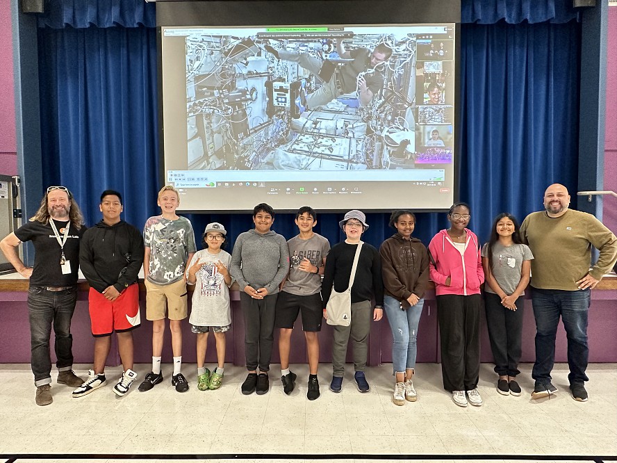9 Zero Robotics students and 2 adult advisors stand before a screen telecast of the ISS as an astronaut tests the team's code