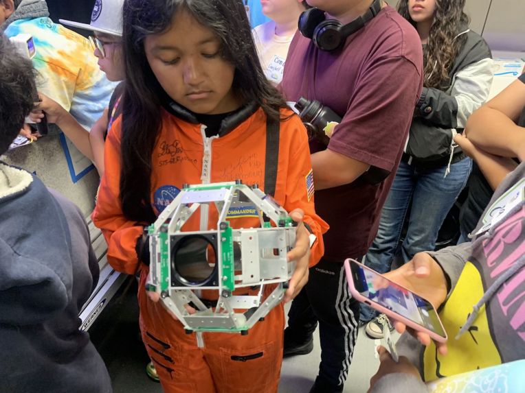 girl in orange NASA space suit holds the Astrobee robot as others look on